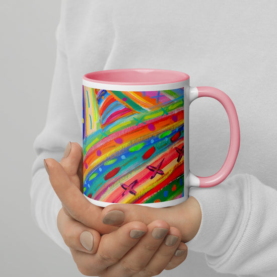 A person holding My Favourite Colour is Rainbows The Rainbow Connection Mug, featuring vibrant swirls and dots with a pink handle and rim. Theyre wearing a light gray long-sleeve shirt.