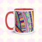 The Rainbow Connection Mug by My Favourite Colour is Rainbow features a vibrant abstract design with stripes, dots, and triangles in bright blue, pink, yellow, and green. The mug features a red interior and handle and is shown against a light decorative background.