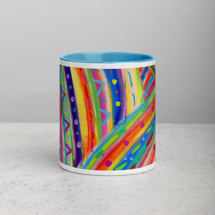 Front view of the Rainbow Connection Mug by My Favourite Colour is Rainbow, featuring a vibrant abstract design with bright blue, pink, orange, and green stripes, dots, and triangles. It has a light blue interior and rests on a grey surface.