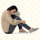 A person wearing a cozy brown sweater and The Rainbow Connection Canvas Sneakers by My Favourite Colour is Rainbow sits on the ground, resting their head on an arm propped against their knee. The background showcases a pattern of pink stars on a light surface.