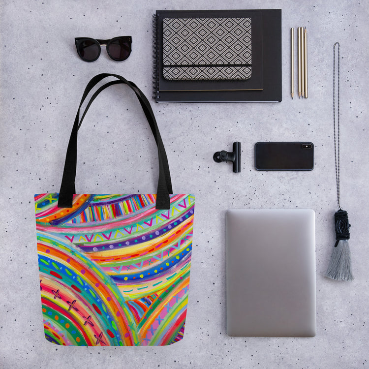 Flat lay of My Favourite Colour is Rainbow's The Rainbow Connection Tote Bag, a laptop, smartphone, black sunglasses, pens, notebooks with geometric designs, and a gray tassel keychain arranged on a speckled gray surface.