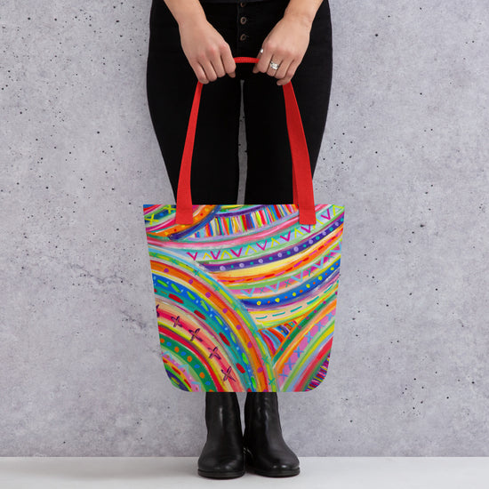 The person holds The Rainbow Connection Tote Bag by My Favourite Colour is Rainbow, with abstract multicoloured swirls and patterns and red handles. They're wearing black pants and boots, standing against a speckled grey wall.