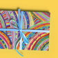 A gift clad in The Rainbow Connection Wrapping Paper from My Favourite Colour is Rainbow, featuring abstract rainbow designs and a light blue ribbon.  Shown on a yellow background.