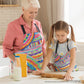 A woman and a young girl in vibrant kitchen attire are baking together. The girl, in a Rainbow Connection Apron by My Favourite Colour is Rainbow, rolls dough on the table with a milk bottle, bowl, and cereal box nearby. They smile warmly in the bright kitchen, enjoying a delightful cooking experience. The apron features vibrant abstract patterns with swirls, dots, and crosses in pink, blue, yellow, and green 