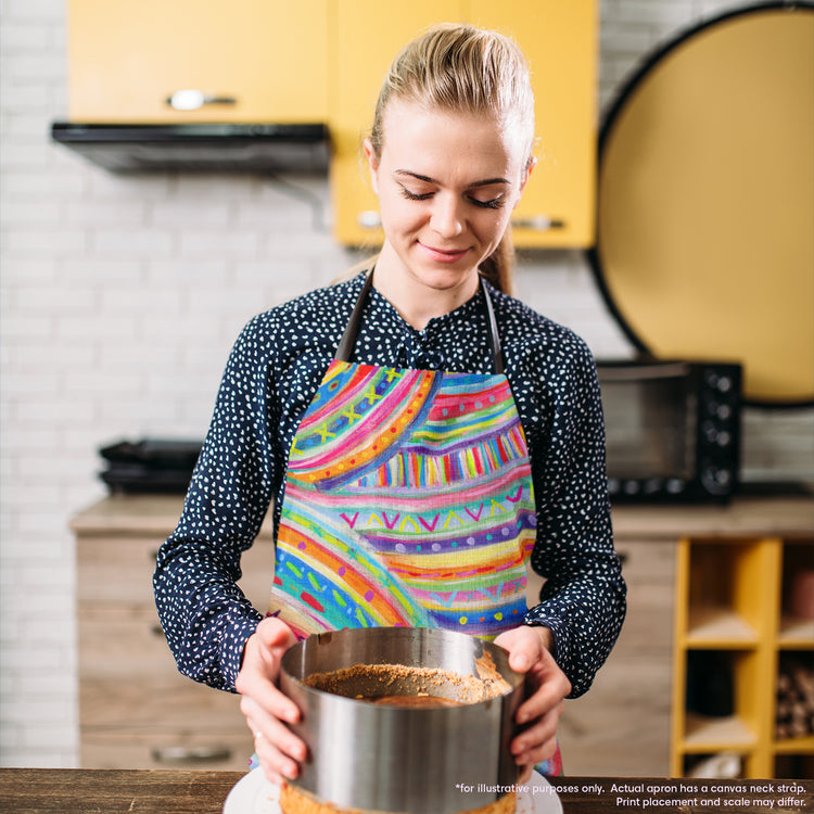 A person wearing the Rainbow Connection Apron by My Favourite Colour is Rainbow holds a round metal baking tin filled with crumb crust. They are in a kitchen with yellow cabinets, a microwave, and a stove in the background, showcasing their cooking expertise.