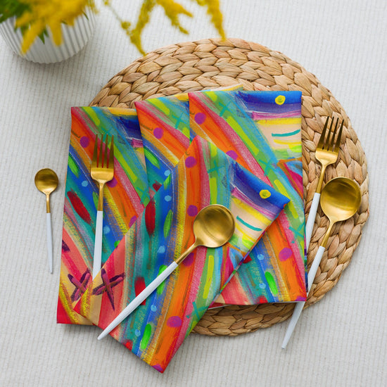 Three vibrant napkins from the Rainbow Connection Cloth Napkin Set by My Favourite Colour is Rainbow highlight a round woven placemat. Theyre paired with gold-colored cutlery, including two spoons and forks, for a chic dining look. Yellow-flowered plants add a touch from the corner.