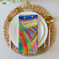 Enhance your dining experience with a white plate on a woven placemat, golden cutlery, and the Mixed Set of 4 Art Napkins by My Favourite Colour is Rainbow, featuring vibrant blues, oranges, and pinks like abstract paintings.