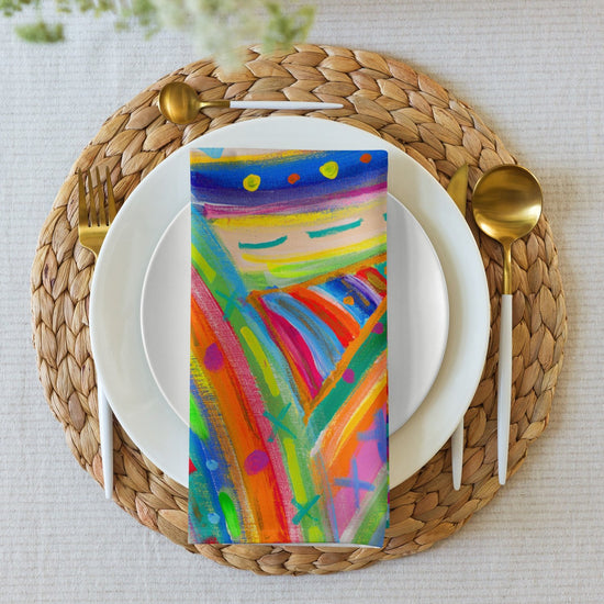 Enhance your dining experience with a white plate on a woven placemat, golden cutlery, and the Mixed Set of 4 Art Napkins by My Favourite Colour is Rainbow, featuring vibrant blues, oranges, and pinks like abstract paintings.