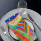 A plate showcases a vibrant napkin from The Rainbow Connection set by My Favourite Colour is Rainbow, nestled between a fork and knife. Above, a clear wine glass sits on a woven placemat, adding elegance to the dark table.