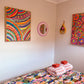 In a colorful bedroom, a bed boasts a vibrant floral bedspread and two rolled towels. The walls feature The Rainbow Connection Framed Original Painting by My Store and another abstract art piece, alongside a small round mirror. A decorative wooden item adds to the rooms cozy, artistic feel.