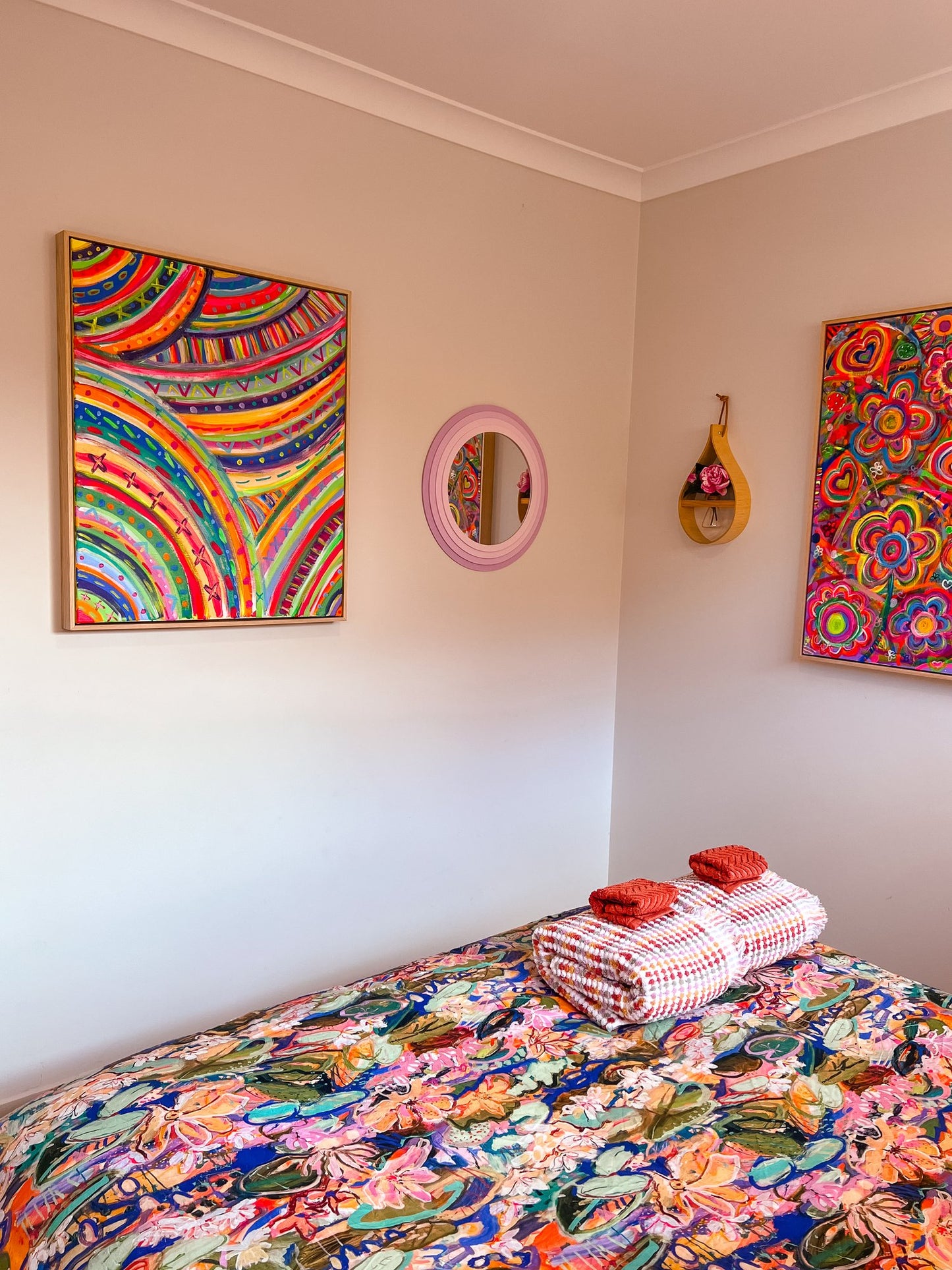 In a colorful bedroom, a bed boasts a vibrant floral bedspread and two rolled towels. The walls feature The Rainbow Connection Framed Original Painting by My Store and another abstract art piece, alongside a small round mirror. A decorative wooden item adds to the rooms cozy, artistic feel.