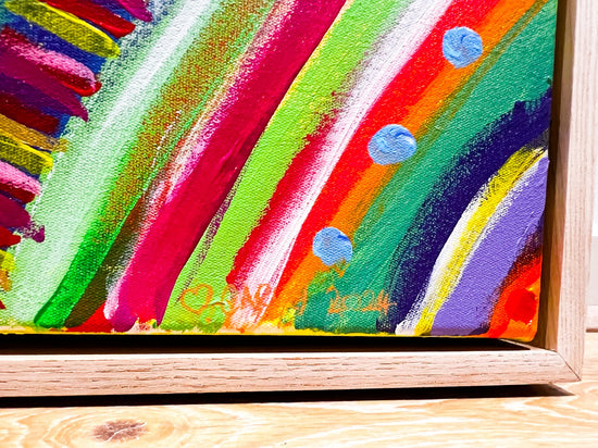 The Rainbow Connection Framed Original Painting by My Store showcases an abstract design with vibrant red, green, orange, and purple strokes plus light blue circles. This acrylic on canvas is professionally framed in a wooden frame with visible wood grain against a wooden background.