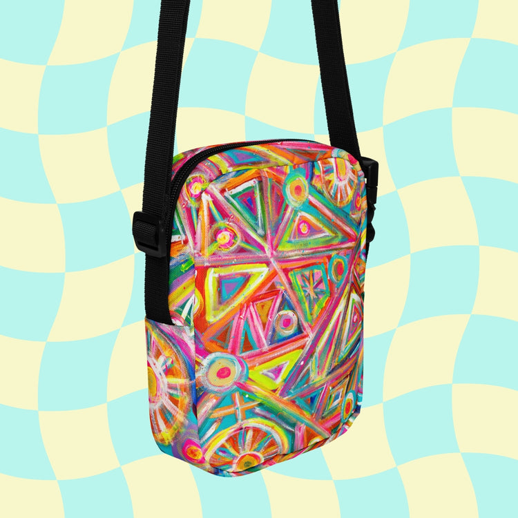 The Tropical Geometry Mini Crossbody Bag by My Favourite Colour is Rainbow showcases colorful triangles and circles in red, pink, green, and yellow. It features a black strap for hands-free use and stands out against a light blue and cream checkered background.