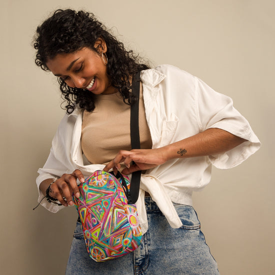 A smiling woman with curly hair, in a white shirt and jeans, opens the My Favourite Colour is Rainbow Tropical Geometry Mini Crossbody Bag over her shoulder for hands-free convenience against a plain background.