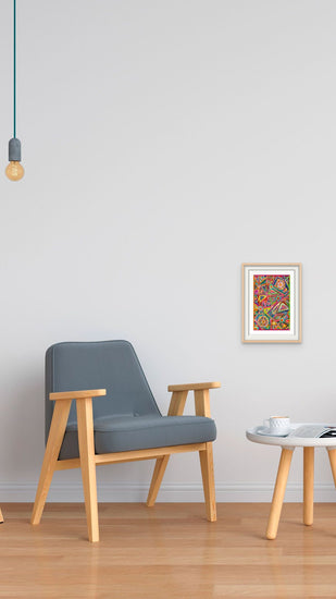 A minimalist room features a gray armchair with a light wood frame, a small round white table holding a cup, and My Stores Tropical Geometry Fine Art Print on the wall. A single hanging light bulb casts gentle shadows on the polished hardwood floor.