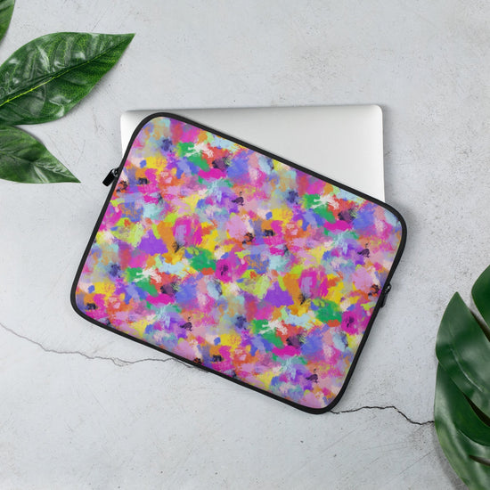 A laptop peeks out of My Favourite Colour is Rainbow's 13 inch Watercolour Rainbow Laptop Sleeve, decorated with abstract splashes of pink, purple, yellow, green, and blue. It rests on a light gray surface surrounded by lush green leaves.