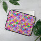 A laptop peeks out of My Favourite Colour is Rainbow's 15 inch Watercolour Rainbow Laptop Sleeve, decorated with abstract splashes of pink, purple, yellow, green, and blue. It rests on a light gray surface surrounded by lush green leaves.