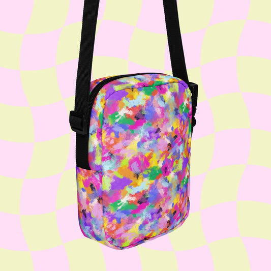 The Watercolour Rainbow Mini Crossbody Bag by My Favourite Colour is Rainbow showcases a vibrant print with abstract paint splatters in pink, yellow, green, blue, and purple. It features an adjustable black strap and pastel checkered background.