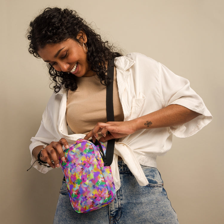 A person with curly hair, in a white shirt and jeans, smiles while holding the My Favourite Colour is Rainbow Watercolour Rainbow Mini Crossbody Bag; it showcases a vibrant print and adjustable straps against a beige backdrop.