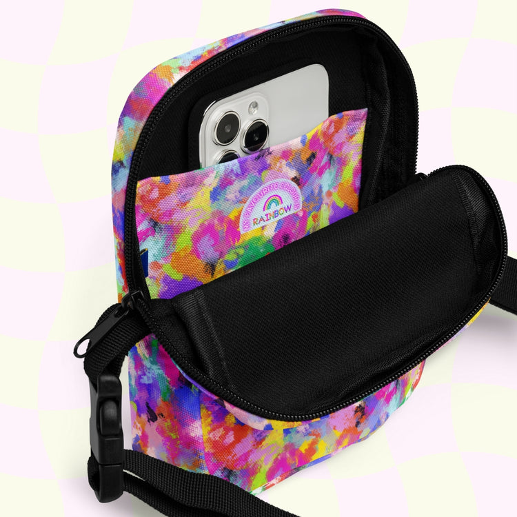 The Watercolour Rainbow Mini Crossbody Bag by My Favourite Colour is Rainbow has a colorful design with an open front pocket showing a white smartphone. It features comfy black adjustable straps and the pocket has a Rainbows patch. The background showcases a pink and yellow checkered pattern.