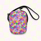 The Watercolour Rainbow Mini Crossbody Bag by My Favourite Colour is Rainbow showcases vibrant pink, blue, yellow, and green splashes on a pastel checkered background. It features adjustable straps and a sleek black zipper on top.