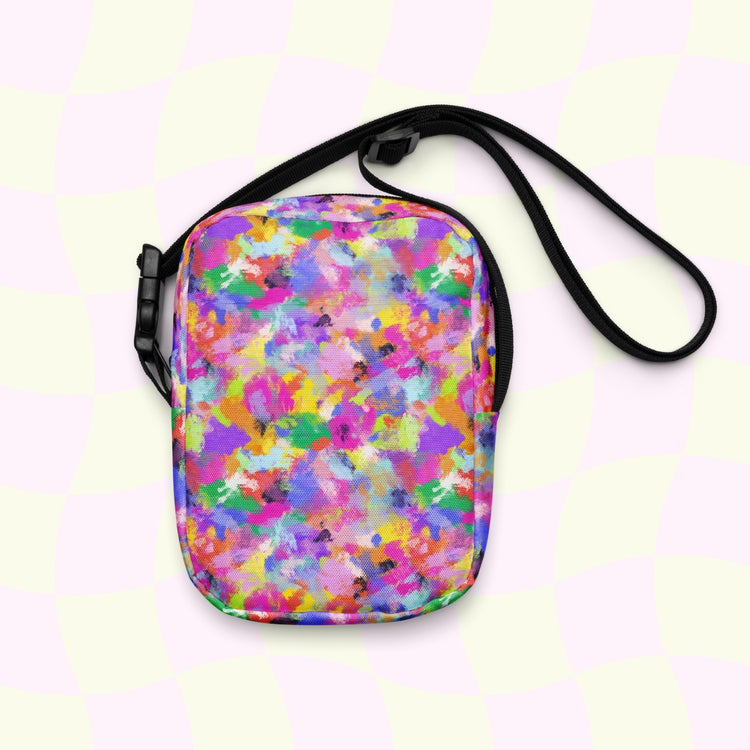 The Watercolour Rainbow Mini Crossbody Bag by My Favourite Colour is Rainbow showcases vibrant pink, blue, yellow, and green splashes on a pastel checkered background. It features adjustable straps and a sleek black zipper on top.