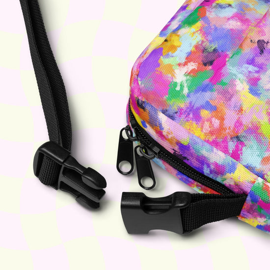 Close-up of the Watercolour Rainbow Mini Crossbody Bag, by My Favourite Colour is Rainbow, features floral patterns, dual black zippers, and an adjustable black strap with a plastic buckle against a white and pale green checkered backdrop.