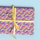 A gift wrapped in My Favourite Colour is Rainbows Watercolour Rainbow Wrapping Paper Sheets features a vibrant abstract design in pink, yellow, green, blue, and white. It is tied with a yellow ribbon bow and set against a light blue background.