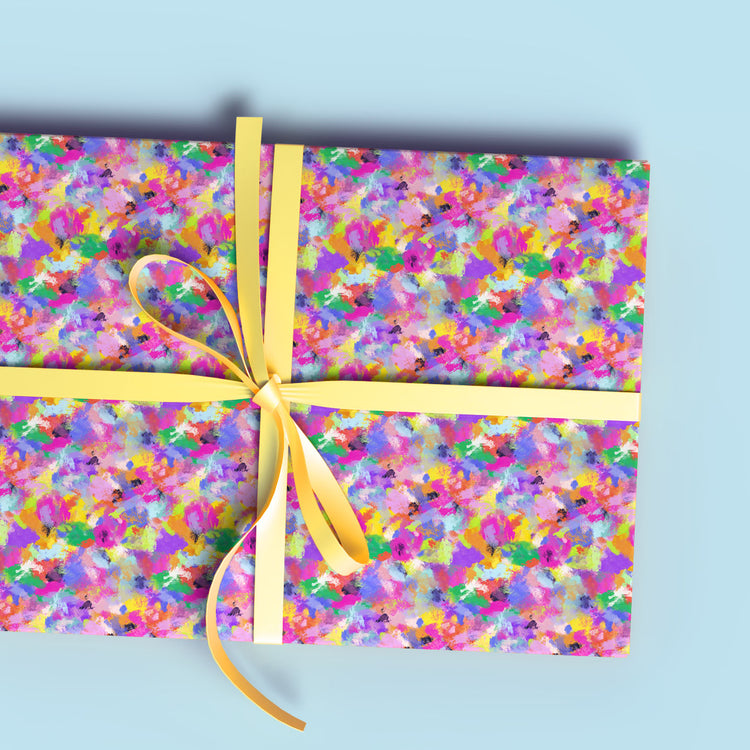 A gift wrapped in My Favourite Colour is Rainbows Watercolour Rainbow Wrapping Paper Sheets features a vibrant abstract design in pink, yellow, green, blue, and white. It is tied with a yellow ribbon bow and set against a light blue background.