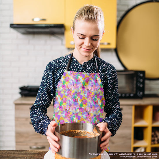 In a kitchen with yellow cabinets and appliances, a woman in a Watercolour Rainbow Apron by My Favourite Colour is Rainbow holds a cake ring filled with dessert.  The apron features an abstract multicoloured pattern with pink, yellow, blue, purple and green.