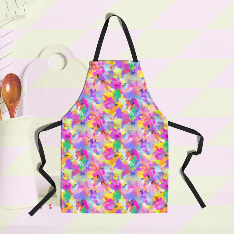 The Watercolour Rainbow Apron by My Favourite Colour is Rainbow features an abstract multicoloured pattern with pink, yellow, blue, purple and green. A white cup utensils holder, wooden spoon and cutting board are in the background.