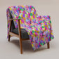 A wooden armchair with gray upholstery is adorned with the Watercolour Rainbow Throw Blanket by My Favourite Colour is Rainbow, featuring an abstract print in pink, purple, yellow, and white. This lively design stands out against the simple light background.