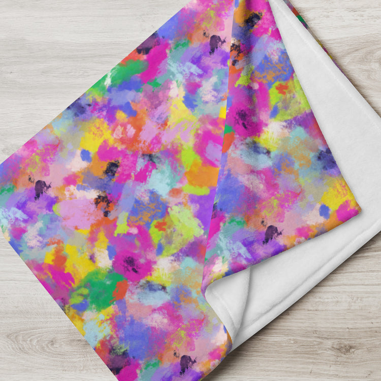 The Watercolour Rainbow Throw Blanket by My Favourite Colour is Rainbow displays a vibrant abstract splatter of purple, pink, yellow, green, blue, and orange on a wooden surface. Its corner is folded to reveal the plain white underside, highlighting the delightful contrast of its soft fabric.