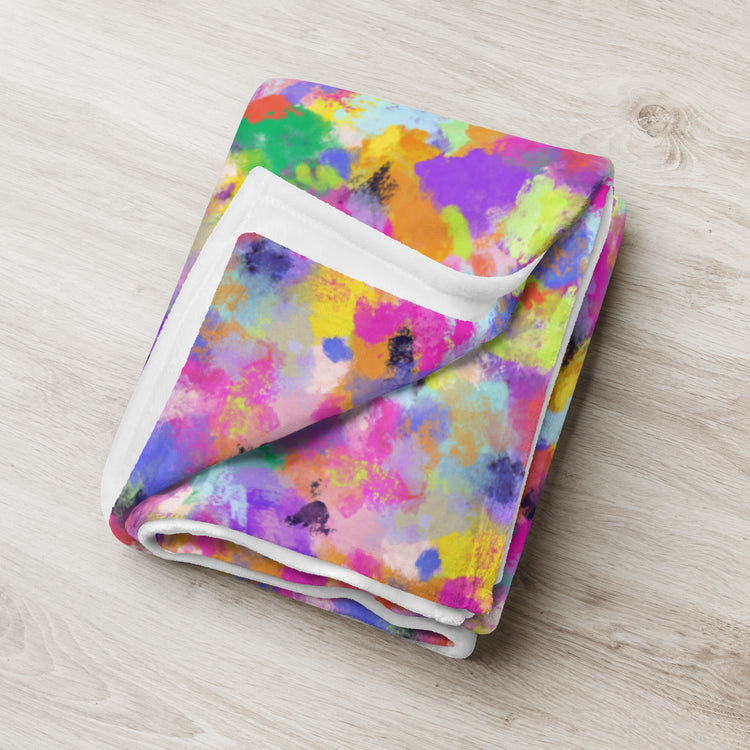 A folded Watercolour Rainbow Throw Blanket by My Favourite Colour is Rainbow showcases a vibrant abstract design with splashes of pink, blue, yellow, green, and purple on a light wooden surface. The soft white-edged fabric enhances the contrast of its multicolored pattern.