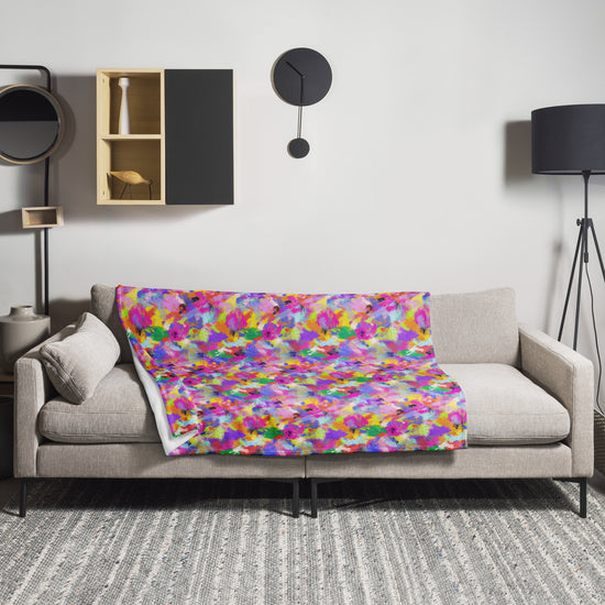 A modern living room showcases a beige sofa with a vibrant Watercolour Rainbow Throw Blanket by My Favourite Colour is Rainbow. The space includes a black floor lamp, wall clock, decor-lined shelves, and a textured gray rug that complements the soft fabric of the blanket.