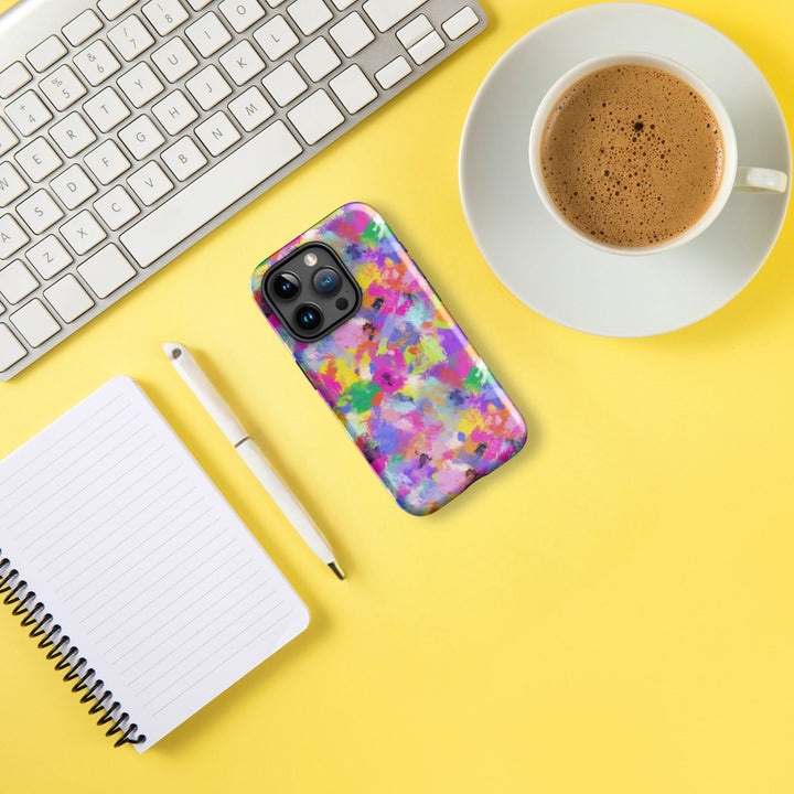 The Watercolour Rainbow MagSafe iPhone Case by My Favourite Colour is Rainbow sits on a smartphone next to a keyboard, coffee cup, open notebook, and pen on a bright yellow surface.