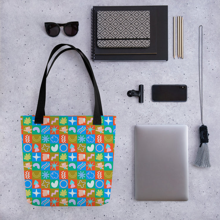 A trendy flat lay displays a spacious Colour Block Tote Bag with abstract shapes by My Favourite Colour is Rainbow, alongside a laptop, smartphone, sunglasses, patterned notebook, pens, phone lens attachment, and a tassel keychain on a speckled gray surface.