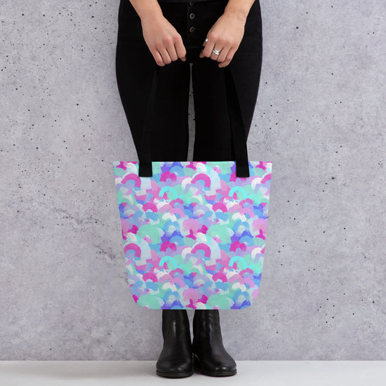 A fashionable person in black jeans and boots carries the spacious Pastel Rainbow Tote Bag from My Favourite Colour is Rainbow, featuring overlapping rainbows in pastel hues. A speckled light grey wall subtly highlights the bags vibrant design.