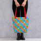 A person in black pants and boots holds the trendy, spacious Colour Block Tote Bag by My Favourite Colour is Rainbow, featuring a stylish pattern of abstract shapes in blue, green, red, and brown and with red straps.