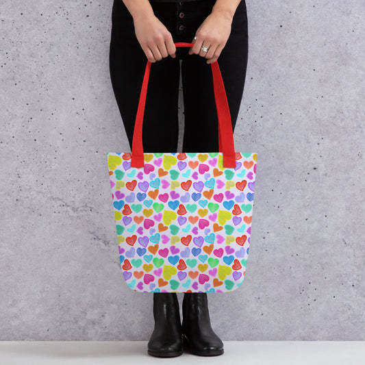 A person holds the Heart Attack Tote Bag by My Favourite Colour is Rainbow, featuring red handles and a vibrant heart design. Paired with black pants and boots against a speckled gray backdrop, this colourful tote adds a pop of flair to the chic look.
