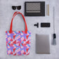 The flat lay features My Favourite Colour is Rainbows Tri the Angle Tote Bag with red handles, perfectly organised with a laptop, smartphone, sunglasses, notebooks, pens, a tassel keychain.