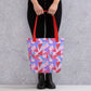 A person holds the Tri the Angle Tote Bag from My Favourite Colour is Rainbow, featuring a colourful geometric design in pink, purple, and red hues with bold red straps. They're wearing black pants and boots against a grey textured wall.