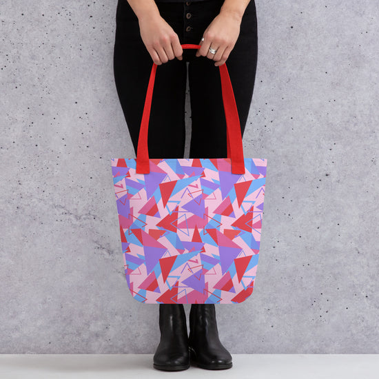 A person holds the Tri the Angle Tote Bag from My Favourite Colour is Rainbow, featuring a colourful geometric design in pink, purple, and red hues with bold red straps. They're wearing black pants and boots against a grey textured wall.