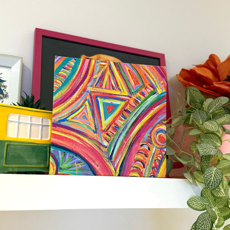 The Karma Kaleidoscope Canvas Artwork by My Favourite Colour is Rainbow showcases intricate geometric neon patterns.  The canvas stands on a white shelf. Nearby, are a variegated leafy plant, an orange flower and other decorative items.