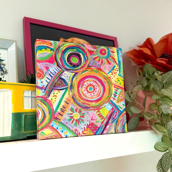 The Summer Sorbet Canvas Artwork by My Favourite Colour is Rainbow, with its vibrant abstract design of circular and spiral patterns, stands a white shelf. Accompanying it are a picture frame, a yellow and green container, an orange flower, and greenery.